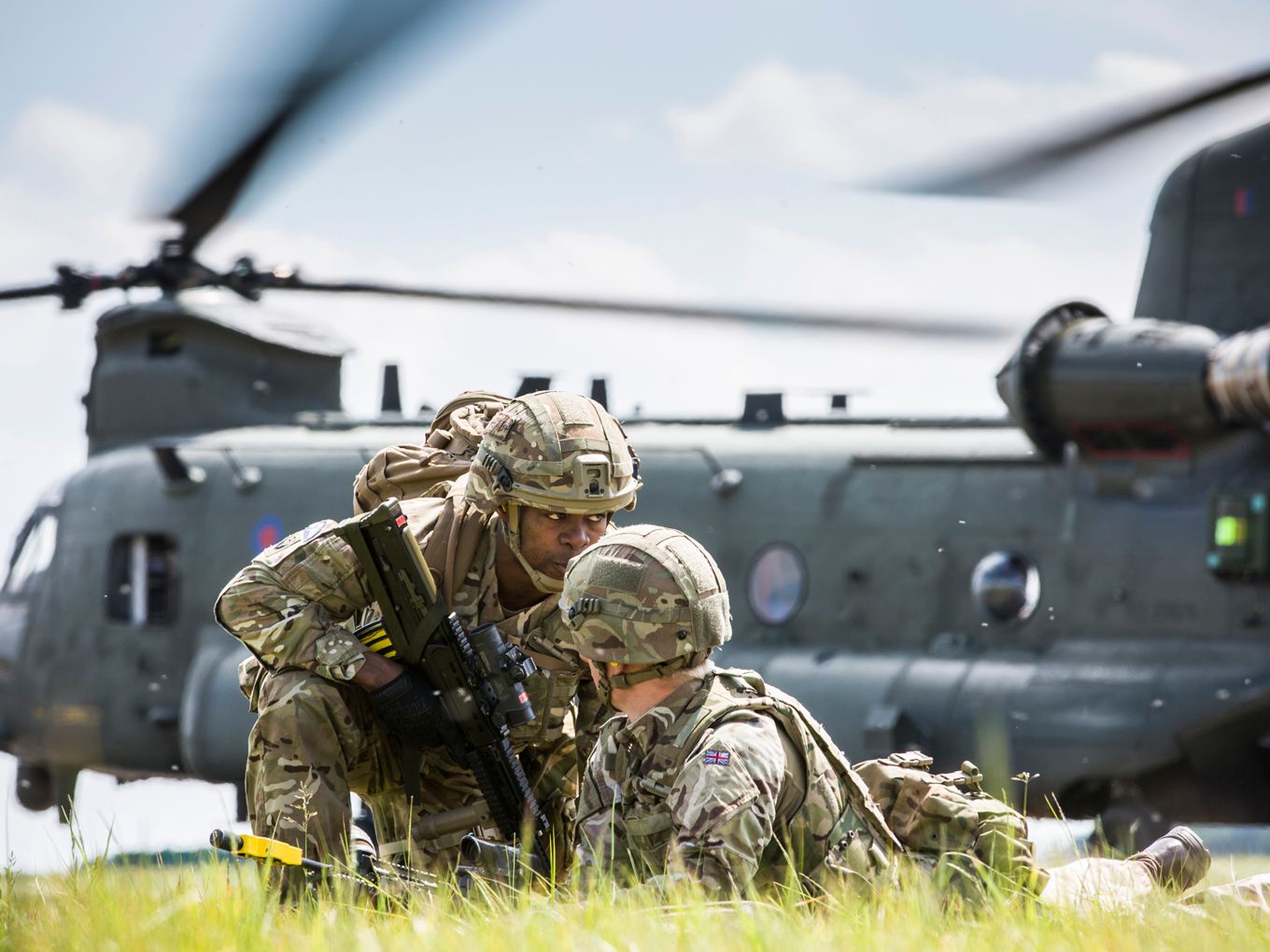Two personnel crouching with a landed Chinook in the background. 