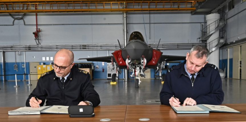Image shows two men signing in books with an F-35 aircraft in the background.