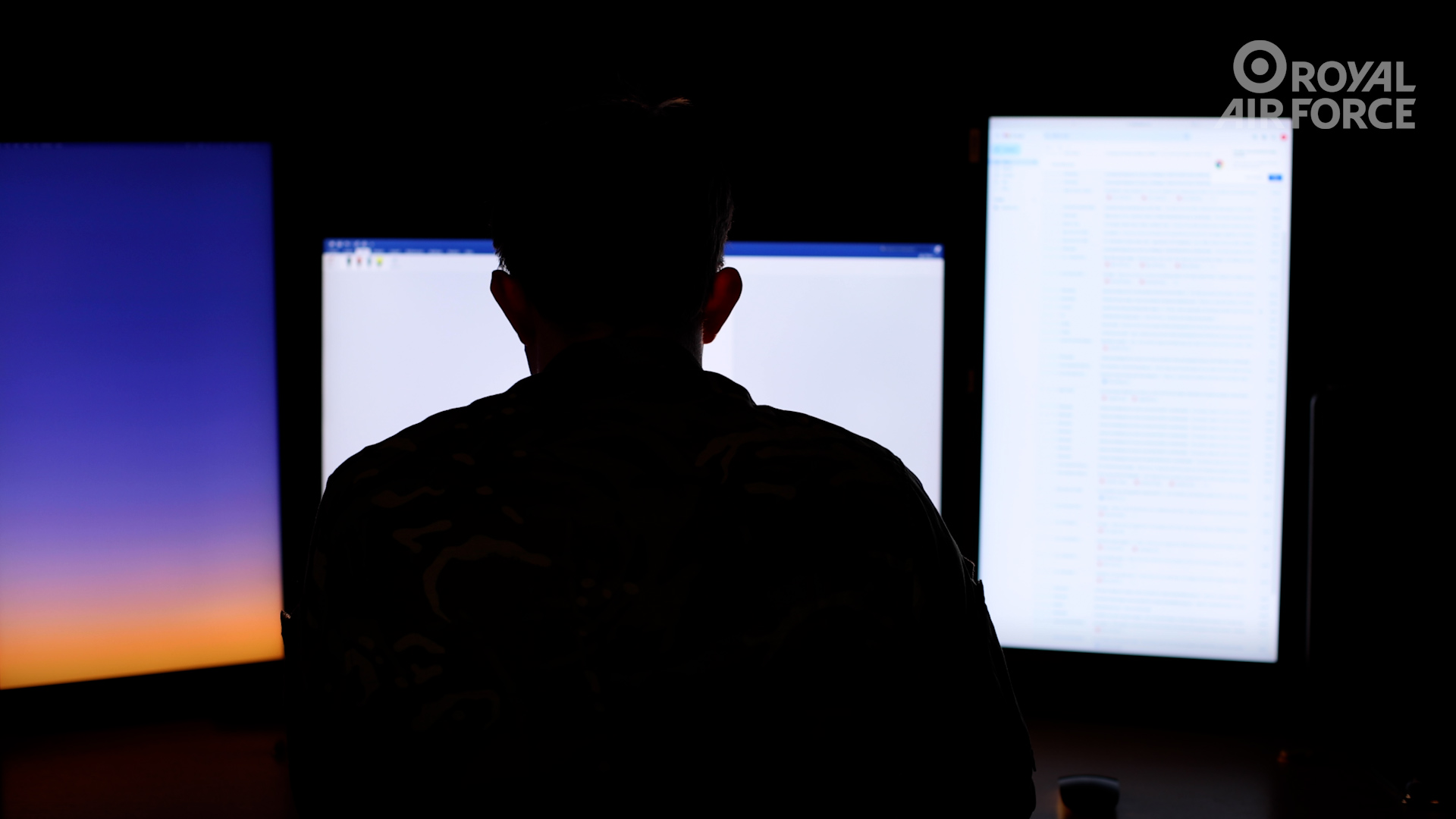 Male silhouette, seated facing three bright monitor screens.