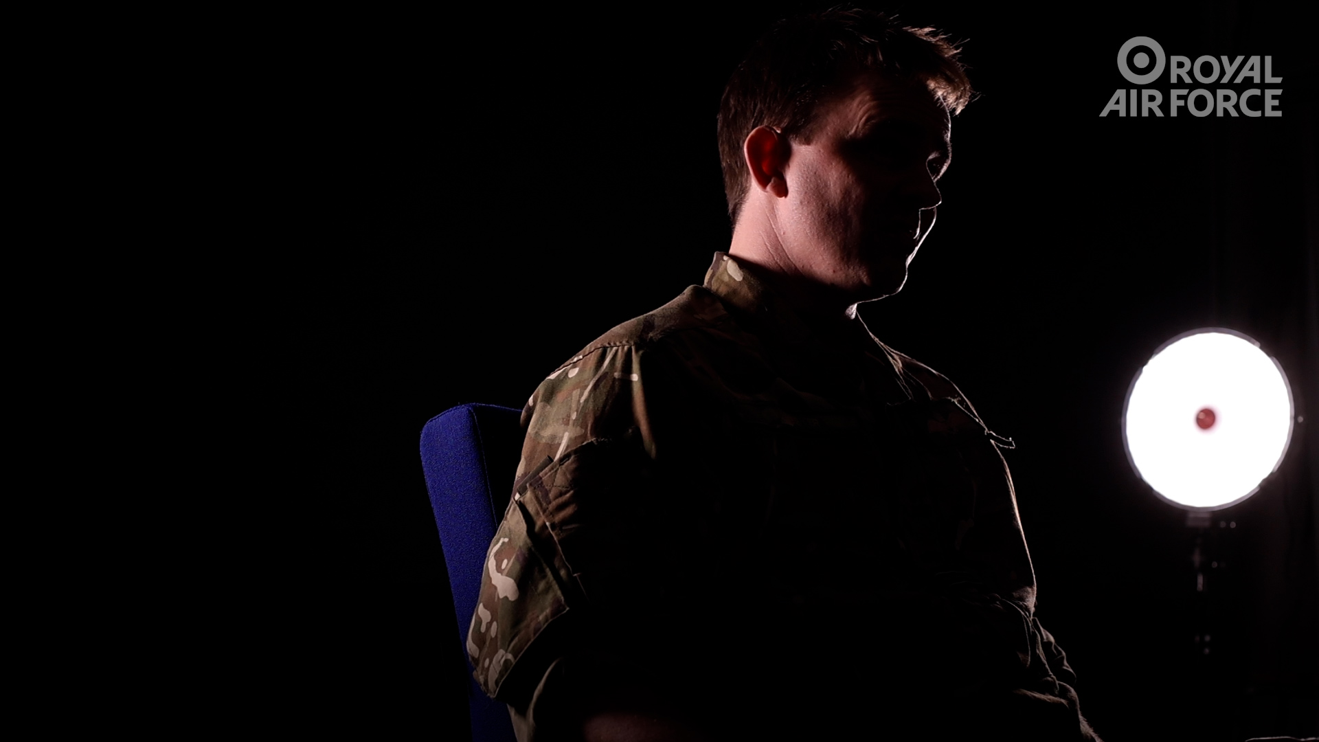 Seated male wearing military uniform in a dimly lit room with a spotlight.