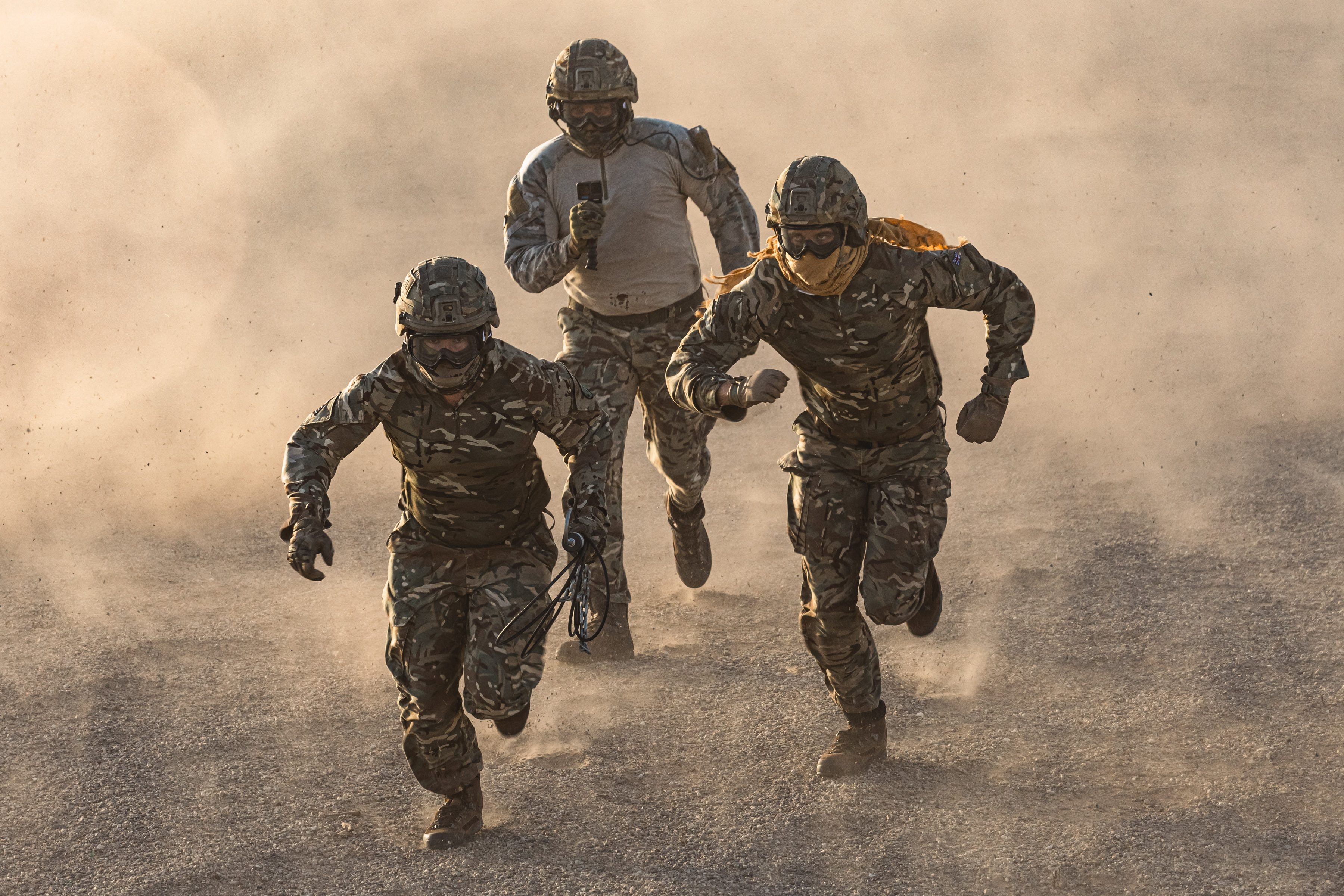 3 Military personnel running across the desert surrounded by dust cloud